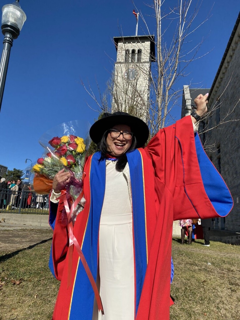 Nel Coloma-Moya in doctoral graduation regalia, holding flowers, with the Grant Hall tower at Queen's University in the background